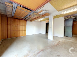 Unfinished basement or workshop space with exposed ceiling joists, wood planks, and metal support straps; concrete floor and white support columns visible.